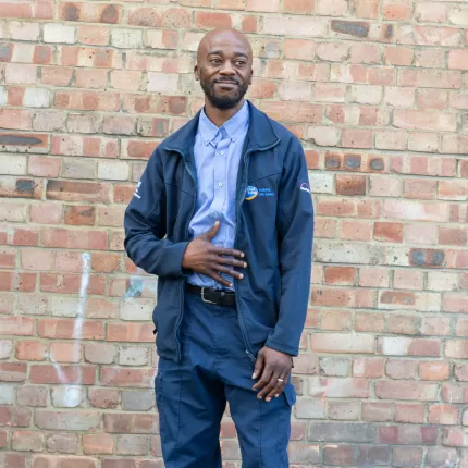 Image of Michael, dressed in a blue workers uniform stood in front of a brick wall